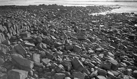 Black and white photograph. Rocks leading back to surf nearly fill this composition. Most of the rocks are rectangular and in a jumble, but some in the distance appear to be stacked in rows.