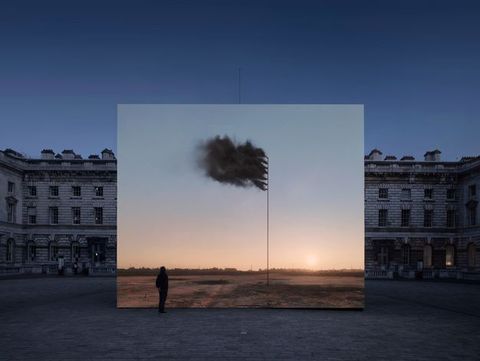 Installation. A digital still on a nearly square screen shows a tall pole stuck in the ground against a flat landscape. Black smoke issuing from seven jets near the top creates a smouldering flag. The screen is set up in front of a long stone building with regular rows of square and rectangular windows.