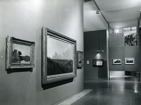 Installation. Black and white photograph of three paintings on a partial wall, with two more hanging on a floating wall across a hall-like space.