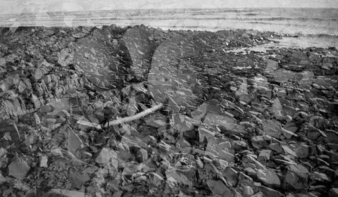 Black and white photograph. Rocks leading back to surf nearly fill this composition. Another image, perhaps of more rocks, is a ghostly echo superimposed on the landscape.