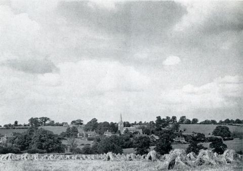 Black and white photograph. A field of haystacks leads to a town of buildings and trees in the lower quarter of this landscape. White clouds march towards us against a pale sky above.