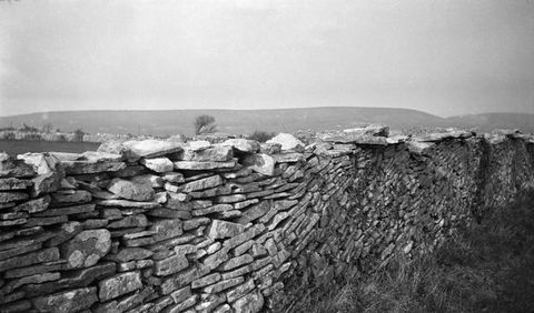 Black and white photograph. Flat rocks stacked to make a wall fall in curves like breaking waves. The wall angles from the lower left corner to the right to cut off our view immediately over the wall. Hills line the horizon in the deep distance.