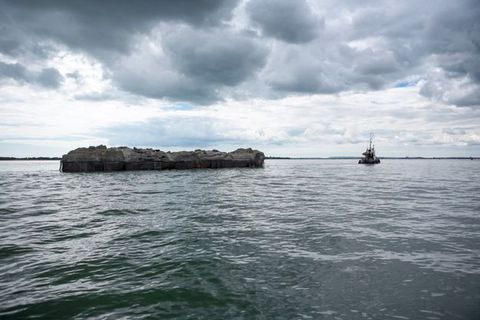 A tugboat pulls what looks like a bit of a rockly island across rippling waters.