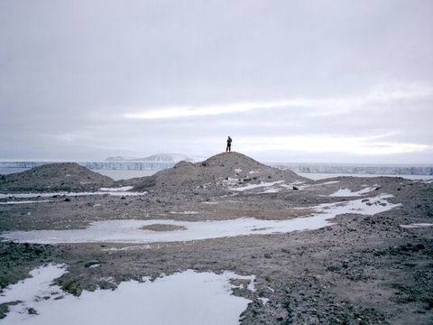 A person is silhouetted against an overcast sky on a low rocky mound. The low areas around the mound are filled in snow.
