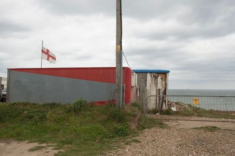 From a sandy coastline we look up at steep cliffs. Buildings perch at the edge are outlined against the overcast sky. Another structure has fallen and crumpled down the cliff face.
