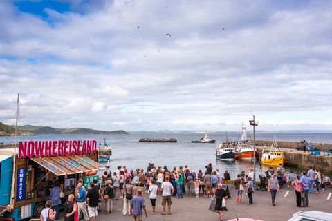 People gather on a concrete walkway along the water’s edge to look out at a rocky island being pulled by a tugboat. A stand to the left is set up. Signs read “Nowhereisland”, “Sign up”, and “Embassy”.