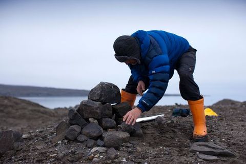 A man wearing a puffy blue coat and orange waders stands on a dark spit of land. He bends forwards and stacks rocks into a dome shape.
