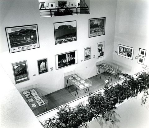 Installation. Black and white photograph overlooking a double-high room hung with eleven posters and works on paper. Three cases along the far wall are filled with open books. A planter in front of us is filled with greenery, which separates us from the space below.